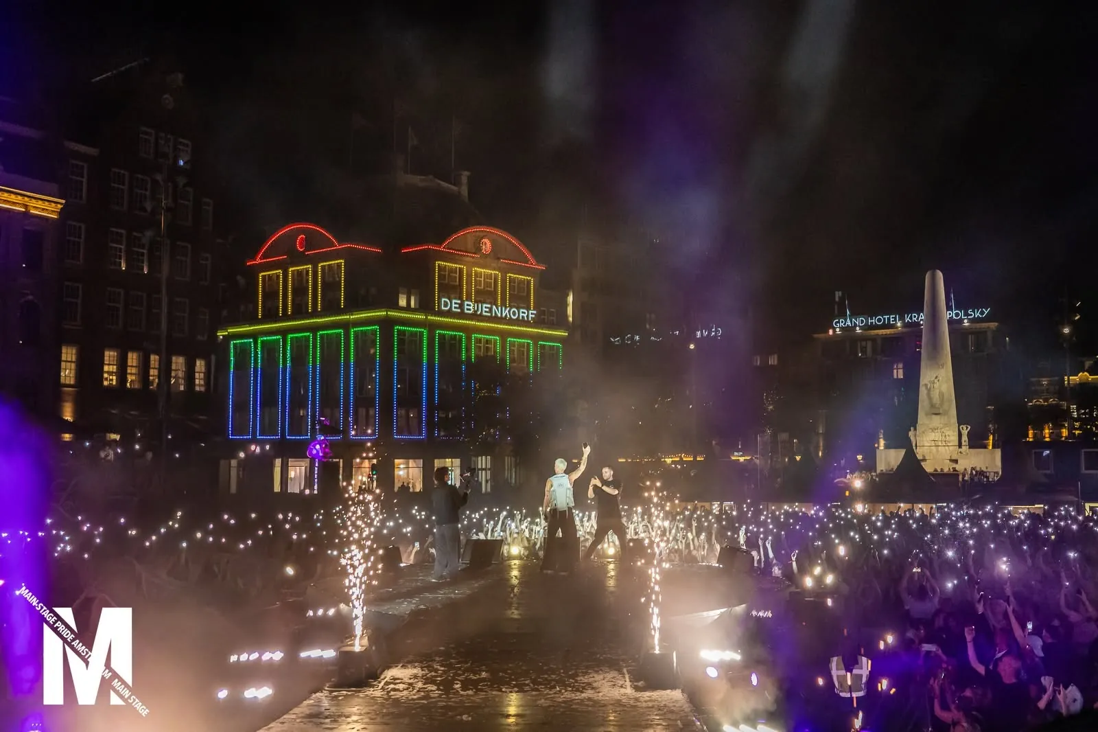 Dam Square at night with a rainbow-lit facade and the crowd of MainStage Pride Amsterdam 2026
